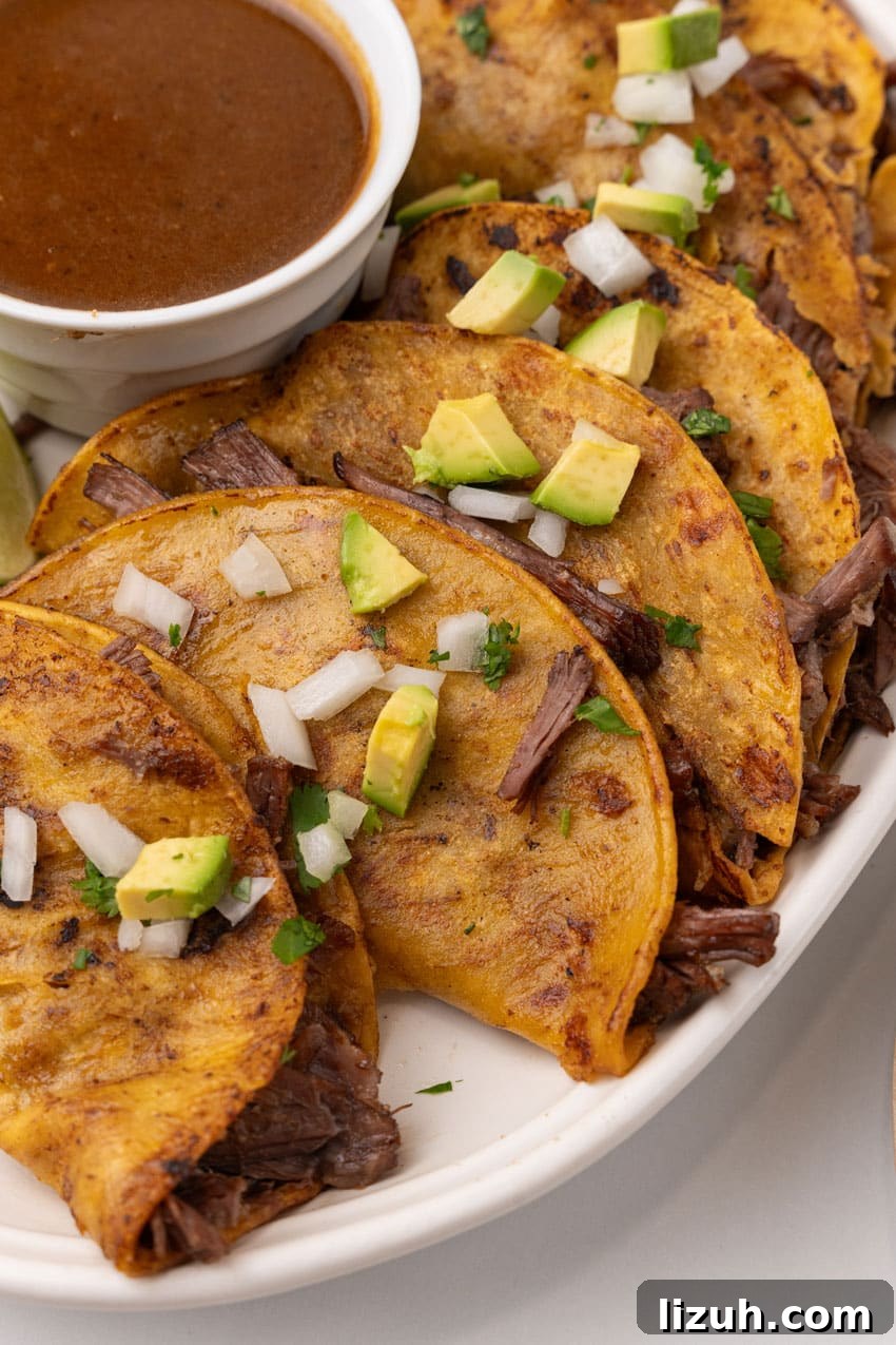 Instant Pot Birria Tacos Recipe — Rich, Juicy Beef Tacos 8 instant pot birria tacos topped with avocado pieces and diced white onion arranged in a row on a white serving platter