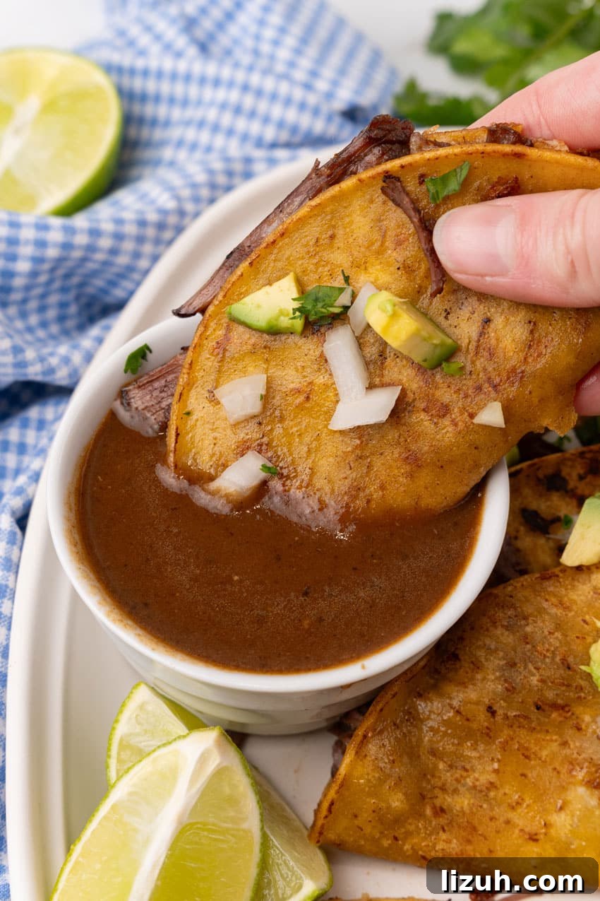 Instant Pot Birria Tacos Recipe — Rich, Juicy Beef Tacos 6 a hand dipping a quesa birria taco in a small white bowl of birria sauce