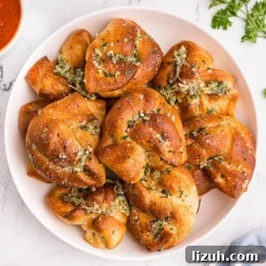 Golden bread knots with garlic and parmesan cheese stacked on a white plate with a small bowl of red sauce.