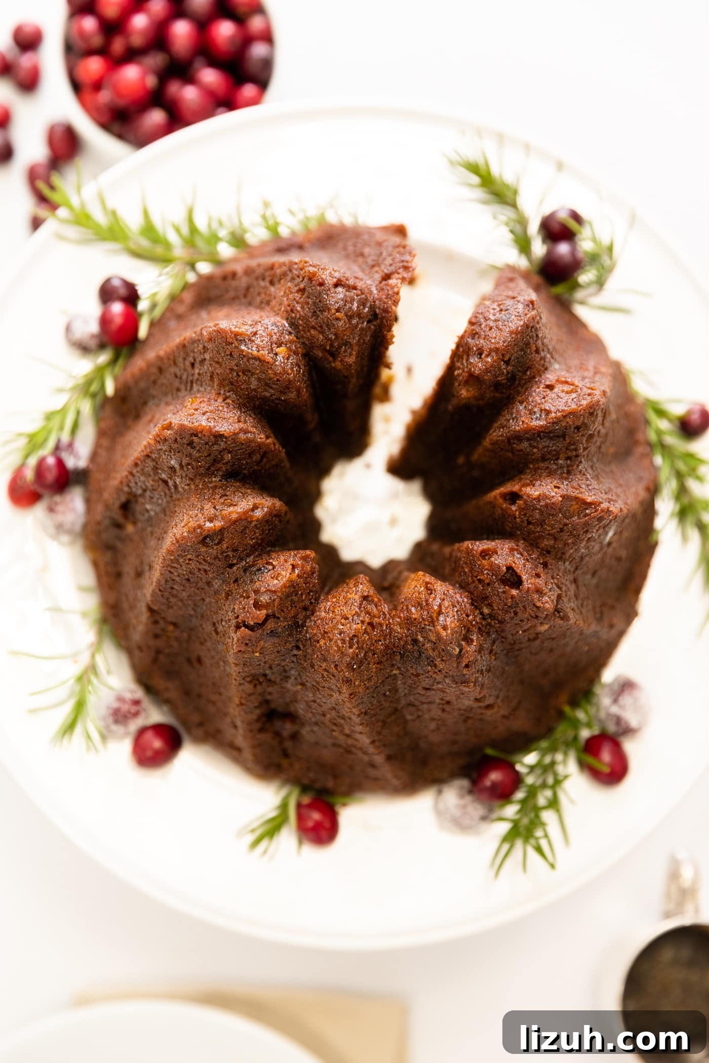 Sticky Date Pudding Cake Recipe with Caramel Sauce 3 Top down view of brown cake made in Bundt pan sitting on white cake plate with cranberries sitting in bowl and piece of cake to side all on white surface