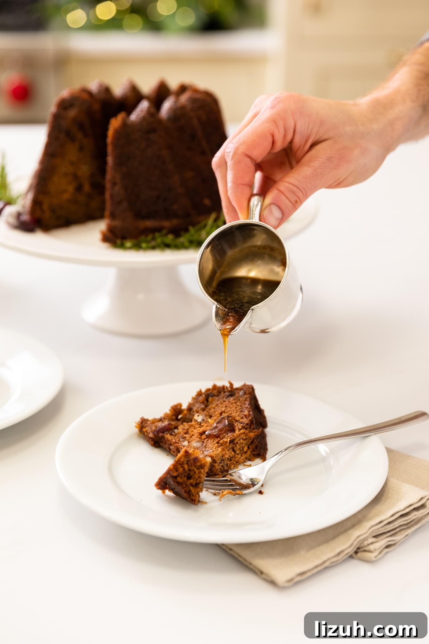 Sticky Date Pudding Cake Recipe with Caramel Sauce 4 Hand pouring sauce onto piece of brown cake sitting on white plate with rest of cake in background all on white surface