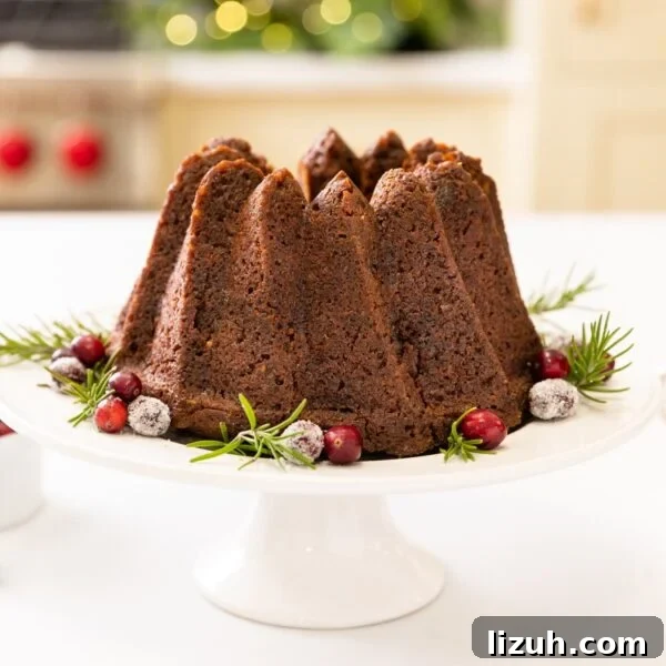 Sticky Date Pudding Cake Recipe with Caramel Sauce 6 Tall brown cake sitting on white cake plate with cranberries sprinkled around all on white surface with oven and cabinetry in background