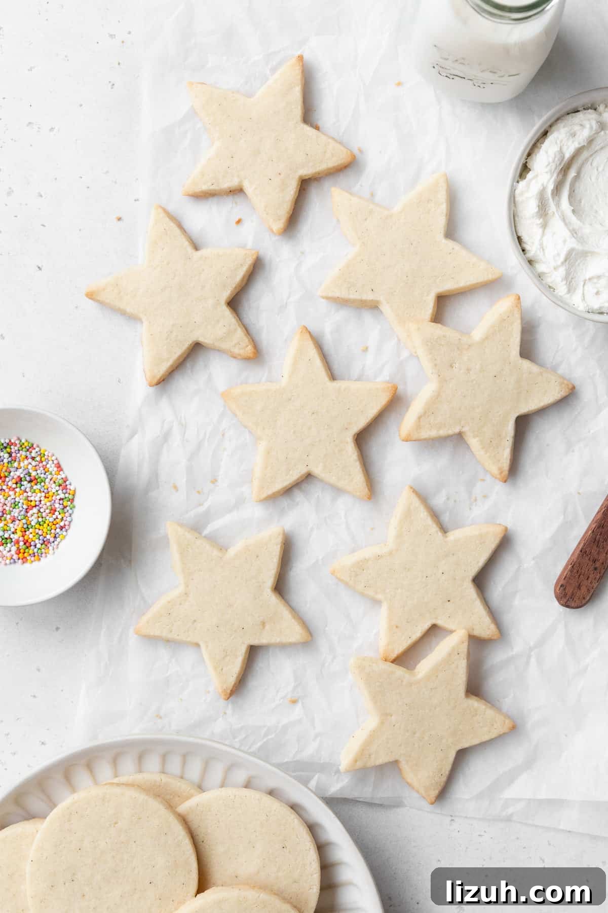 Gluten-Free Sugar Cookie Recipe for Crisp, Chewy Cookies 2 Star shaped gluten free sugar cookies on a white surface, with a small dish of sprinkles, a bowl of frosting, and a plate of circular sugar cookies on a plate surrounding them.