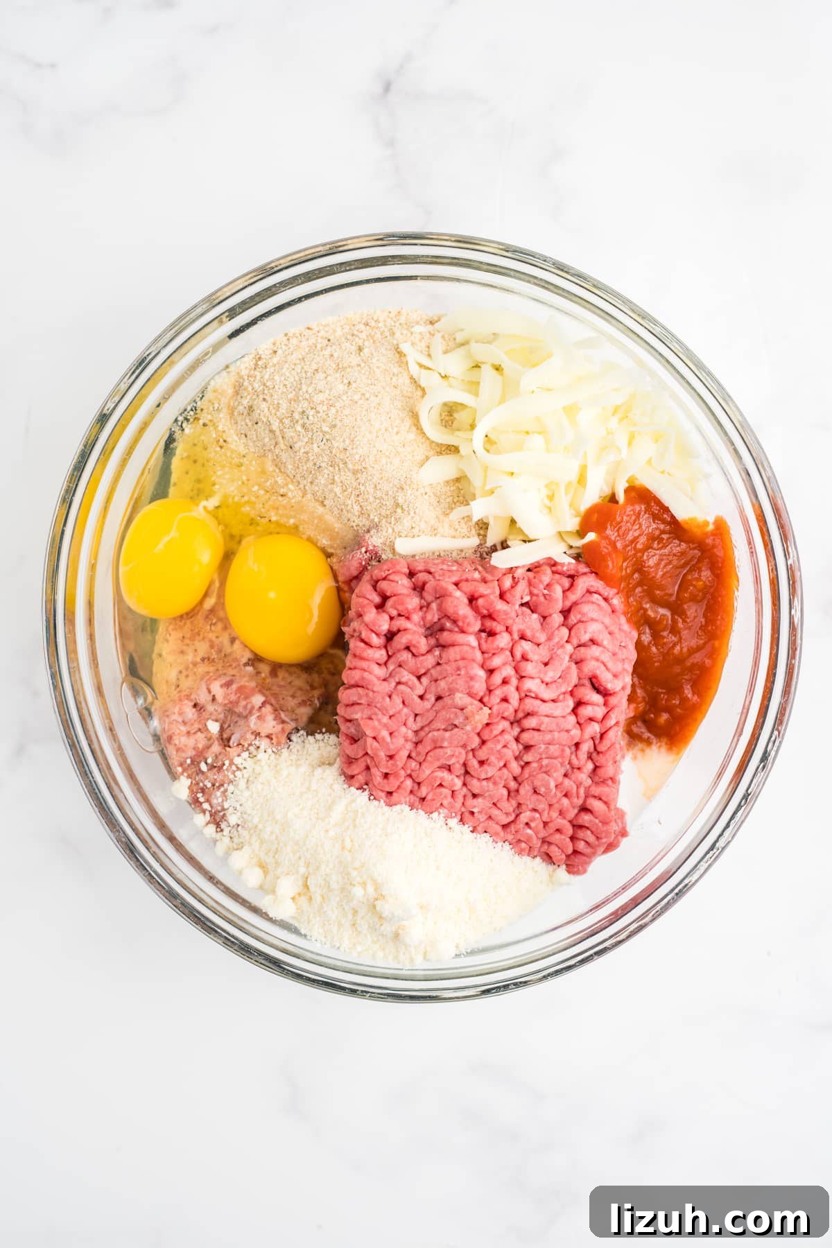 Ground beef, sausage, breadcrumbs, eggs, milk, Parmesan, mozzarella, seasonings, sauteed onion, garlic, and reduced marinara in a bowl.