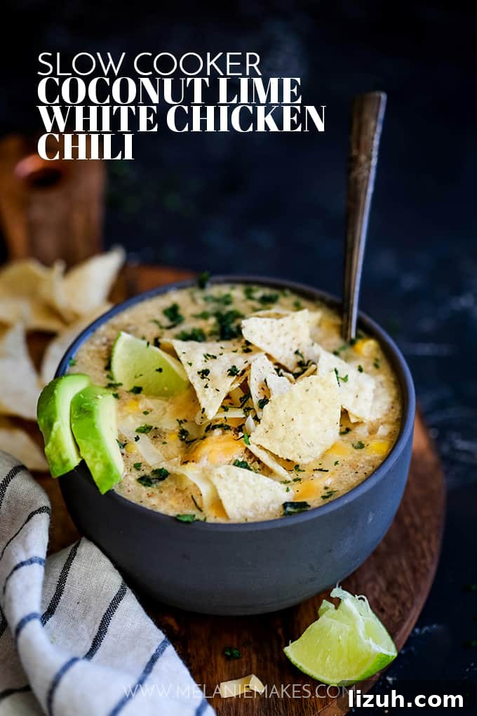 This crazy easy and super flavorful Slow Cooker Coconut Lime White Chicken Chili is a comforting bowl perfect for any day of the week. #slowcooker #crockpot #soup #chili #chicken #chickenrecipes #lime #coconut A bowl of Slow Cooker Coconut Lime White Chicken Chili sits on a wooden surface surrounded by tortilla chips, a striped napkin and a lime wedge.