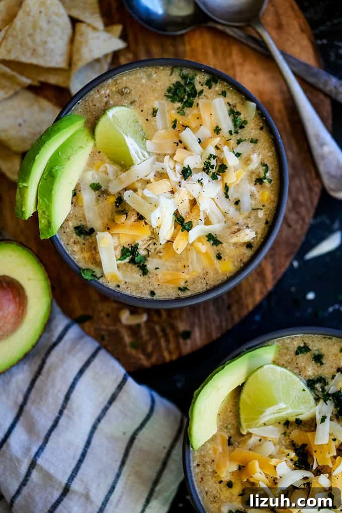 This crazy easy and super flavorful Slow Cooker Coconut Lime White Chicken Chili is a comforting bowl perfect for any day of the week. #slowcooker #crockpot #soup #chili #chicken #chickenrecipes #lime #coconut Two bowls of Slow Cooker Coconut Lime White Chicken Chili sit on a wooden surface surrounded by tortilla chips, a striped napkin, avocado half and two spoons.
