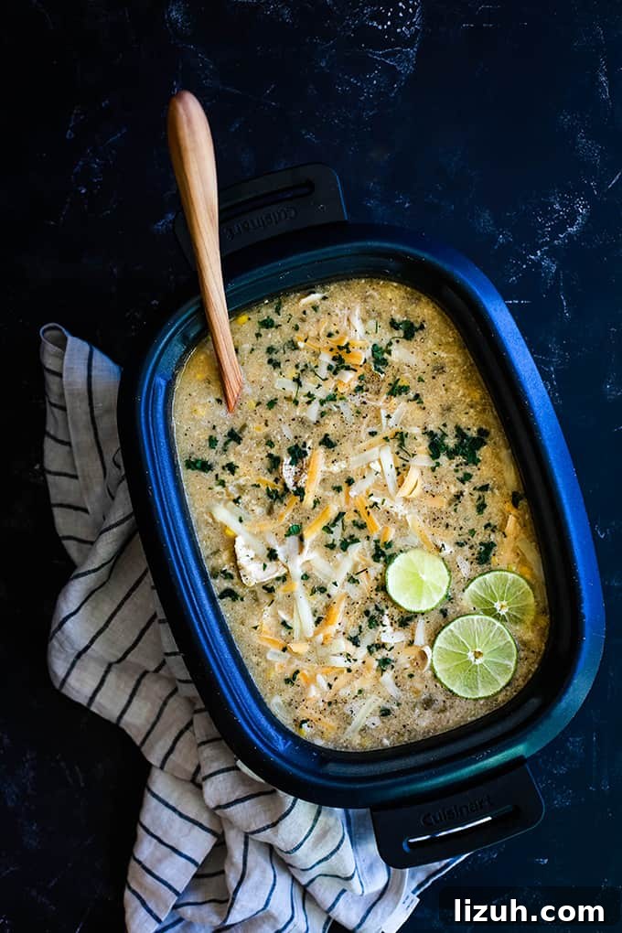 This crazy easy and super flavorful Slow Cooker Coconut Lime White Chicken Chili is a comforting bowl perfect for any day of the week. #slowcooker #crockpot #soup #chili #chicken #chickenrecipes #lime #coconut A slow cooker insert filled with Slow Cooker Coconut Lime White Chicken Chili sits on a dark surface flanked by a striped napkin.
