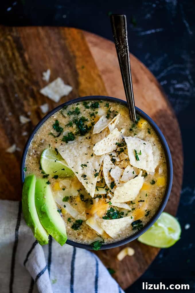 This crazy easy and super flavorful Slow Cooker Coconut Lime White Chicken Chili is a comforting bowl perfect for any day of the week. #slowcooker #crockpot #soup #chili #chicken #chickenrecipes #lime #coconut A bowl of Slow Cooker Coconut Lime White Chicken Chili sits on a wooden surface surrounded by tortilla chips, a striped napkin and a lime wedge.