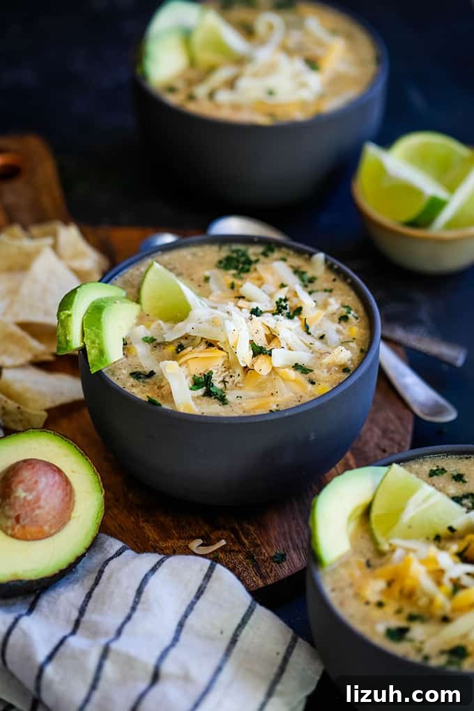 This crazy easy and super flavorful Slow Cooker Coconut Lime White Chicken Chili is a comforting bowl perfect for any day of the week. #slowcooker #crockpot #soup #chili #chicken #chickenrecipes #lime #coconut Three bowls of Slow Cooker Coconut Lime White Chicken Chili sit on a wooden surface surrounded by tortilla chips, an avocado half, a striped napkin and a bowl lime wedges.