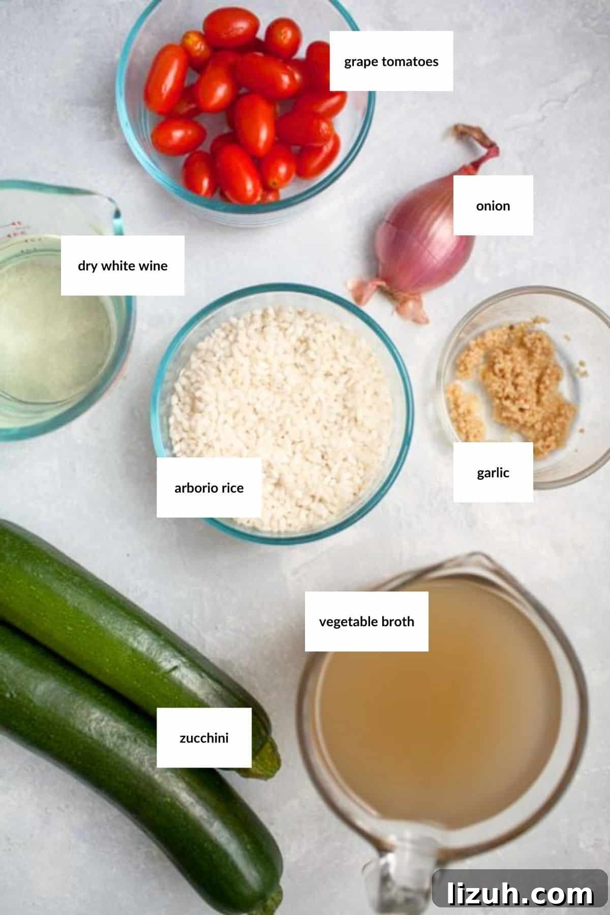 Labeled ingredients for a recipe are arranged on a white surface: grape tomatoes, onion, dry white wine, arborio rice, garlic, zucchini, and vegetable broth.