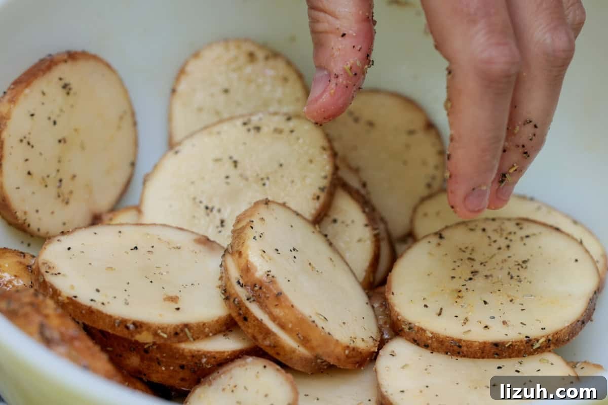 Crispy Grilled Sliced Potatoes Recipe for Summer BBQs 5 Sliced potatoes being hand tossed in olive oil and ingredients in white bowl.