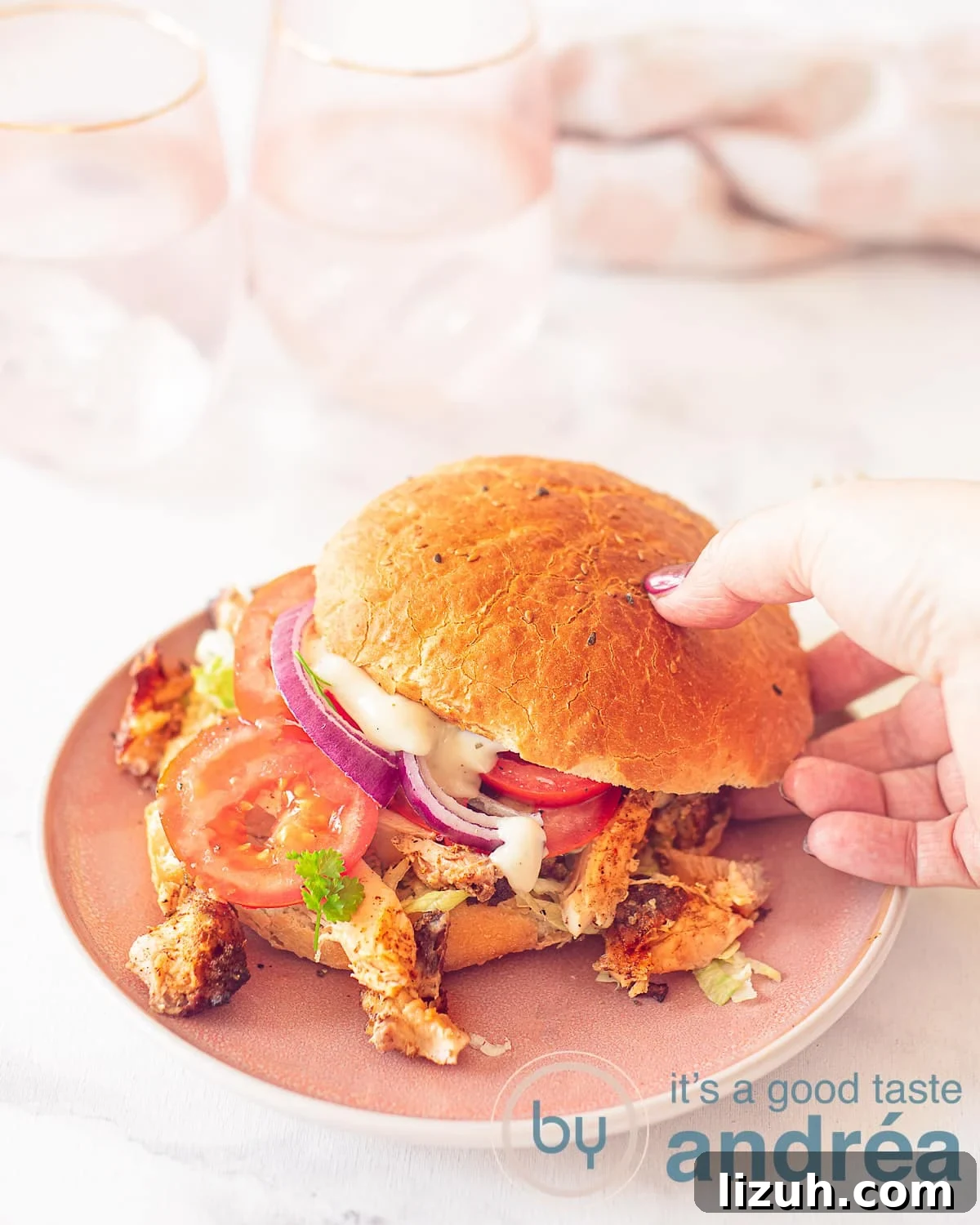 A height photo of a chicken kebab sandwich, raw vegetables on a pink plate. A hand takes the sandwich. A white surface in the background a pink white tea towel