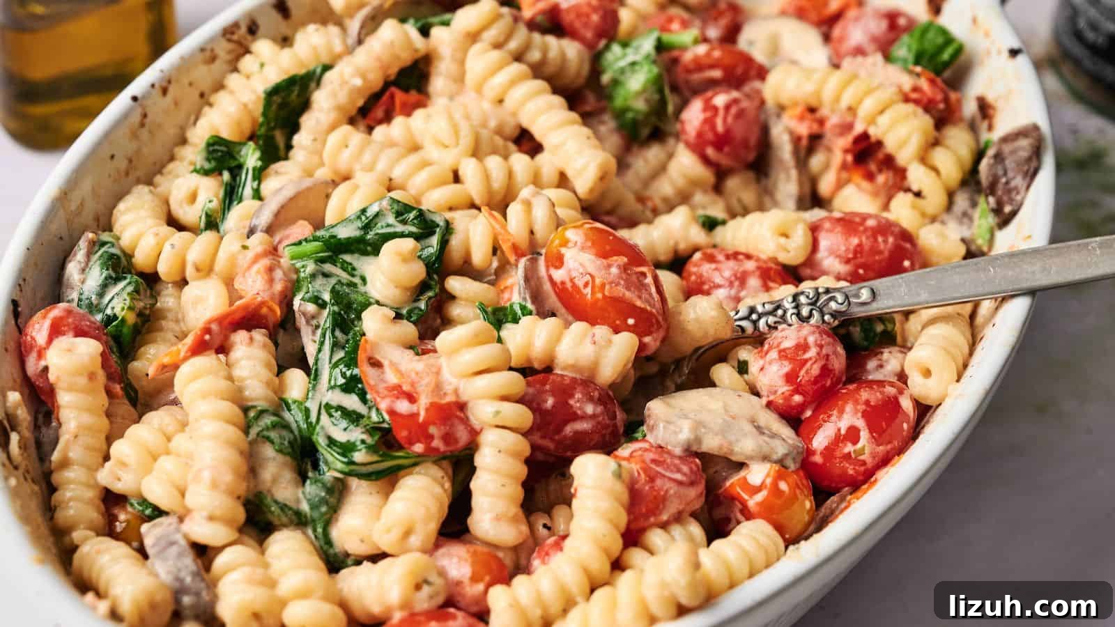 35 Game-Day Recipes to Win Your Season’s Food Lineup 27 A white dish filled with cooked fusilli pasta, cherry tomatoes, spinach, and mushrooms in a creamy sauce, with a silver fork resting inside.