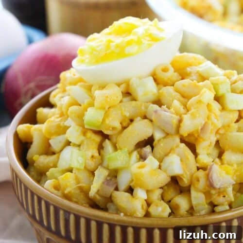 A bowl of Deviled Egg Macaroni Salad with chopped celery and egg, topped with half a boiled egg, sits on a table with an onion and whole eggs in the background.