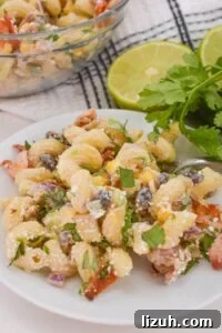A vibrant Mexican Street Corn Pasta Salad is presented, bursting with beans, tomatoes, and fresh herbs alongside lime halves and cilantro on a checked napkin.