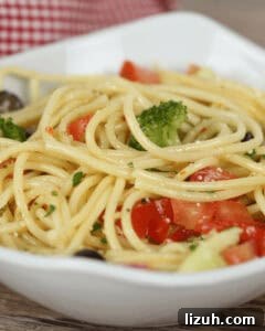 A close-up of a bowl of spaghetti pasta salad mixed with vegetables, including chopped broccoli and tomatoes, garnished with herbs.