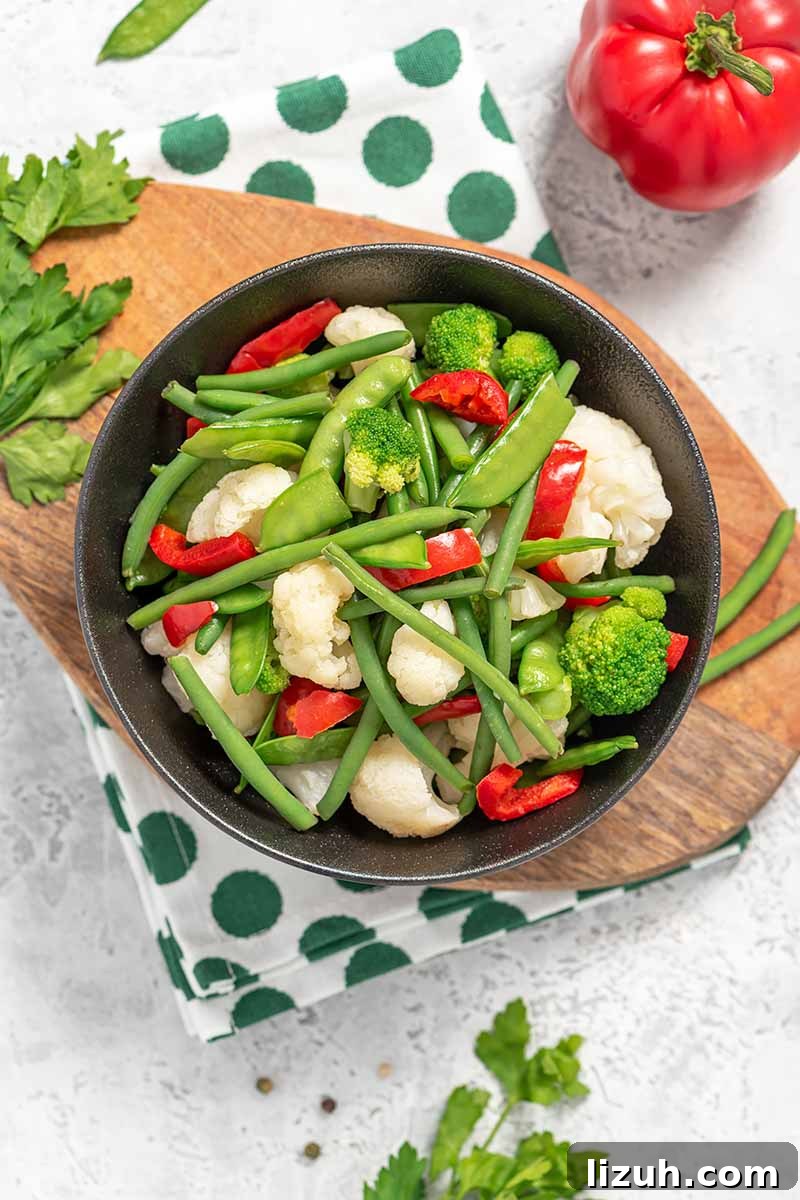 9 Delicious Tricks to Boost Flavor in Steamed Vegetables 3 Vertical top-down image of a large bowl filled with cauliflower, broccoli, beans, peas, and bell peppers on a wooden board with a napkin.
