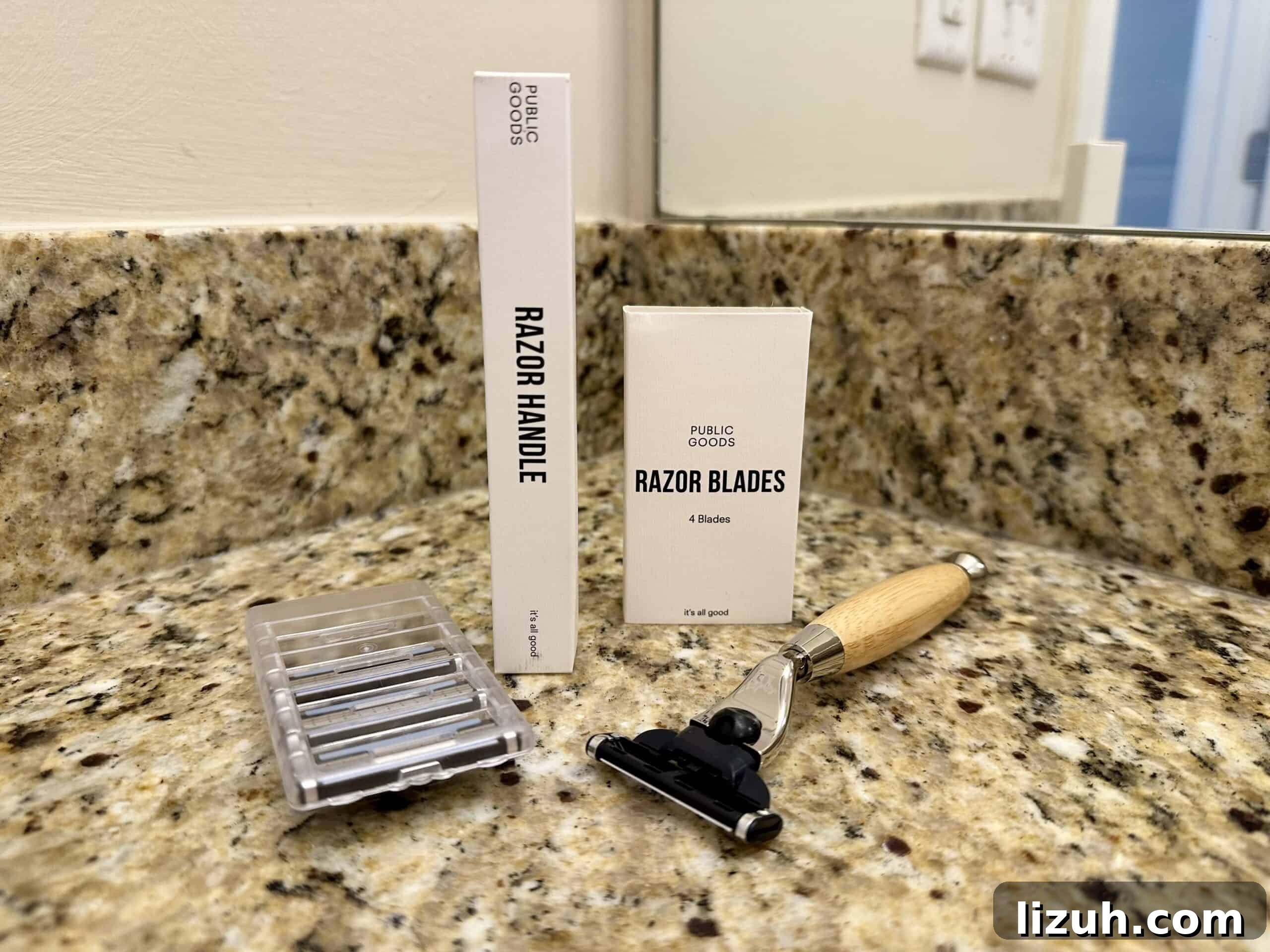 PFAS-Free, Non-Toxic Razors Reviewed: What to Buy in 2026 6 A Public Goods razor set displayed on a granite bathroom countertop, including a wooden-handled razor, a box labeled “Razor Handle,” a box labeled “Razor Blades,” and a plastic case holding replacement blades. A mirror and wall outlet are visible in the background.