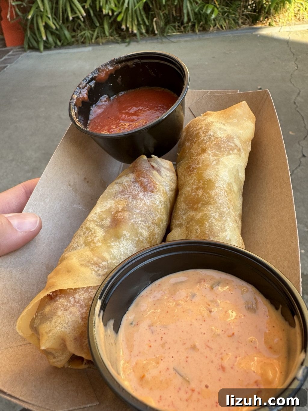 Two cheeseburger spring rolls with a red sauce and a pink sauce in an open cardboard container