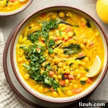 overhead view of a bowl of Golden Orzo Soup with Chickpeas topped with a lemon wedge, green herbs, and nuts.