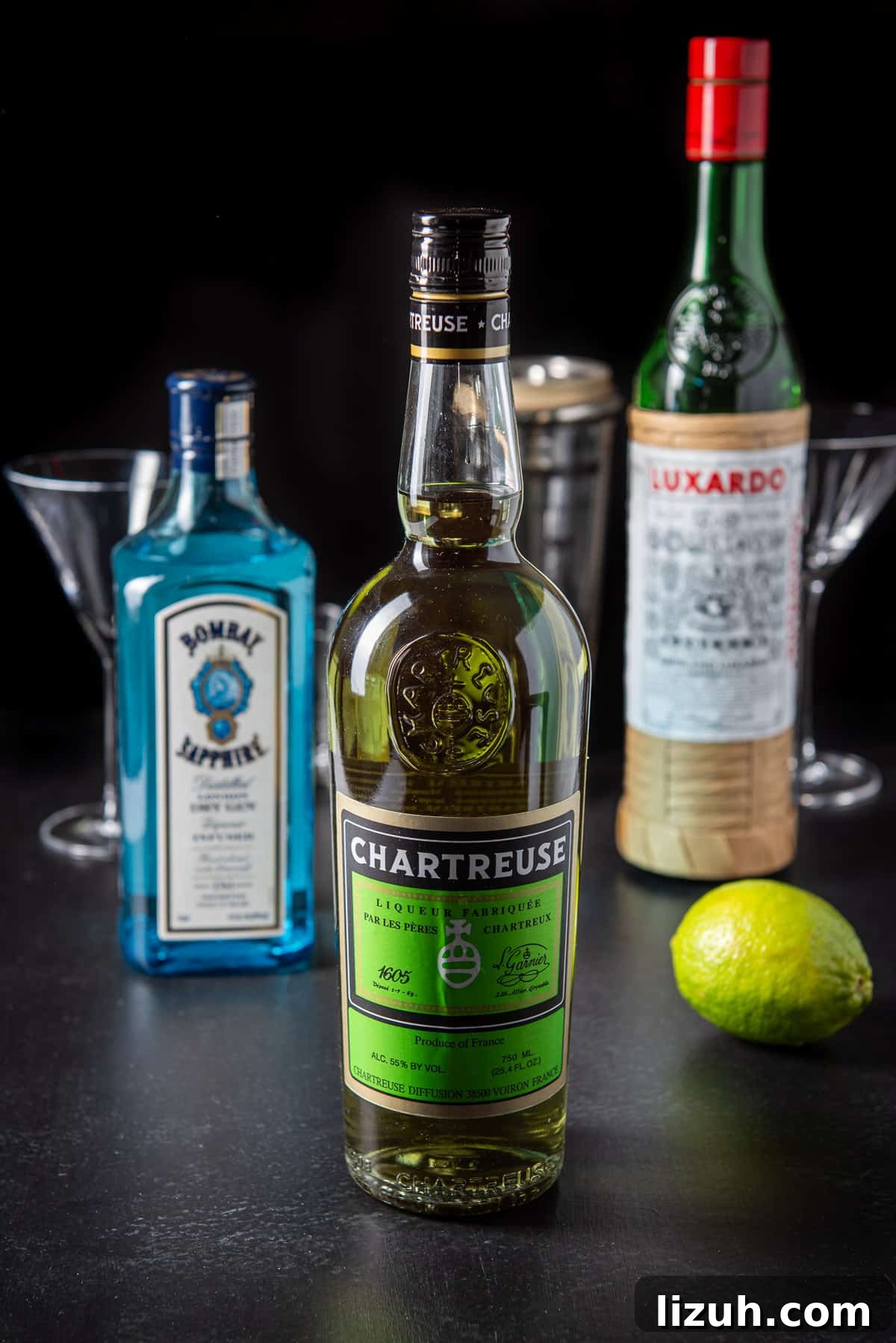 Classic Last Word Cocktail Recipe: How to Mix the Prohibition-Era Drink 4 Bottles of Chartreuse, gin, maraschino cherry and a lime on a table with glasses and the shaker