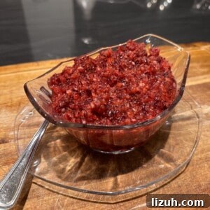 3-Ingredient Cranberry Relish Recipe for Holiday Tables 3 A glass bowl filled with a chunky, red cranberry relish sits on a matching glass plate atop a wooden surface. A metal spoon is placed beside the bowl.