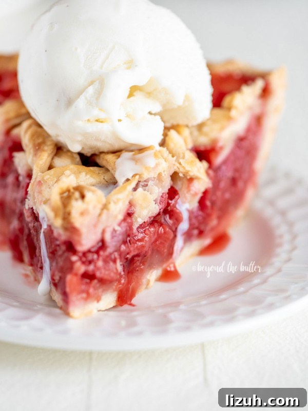 Slice of strawberry rhubarb pie with scoop of vanilla ice cream on dessert plate on white wood table.