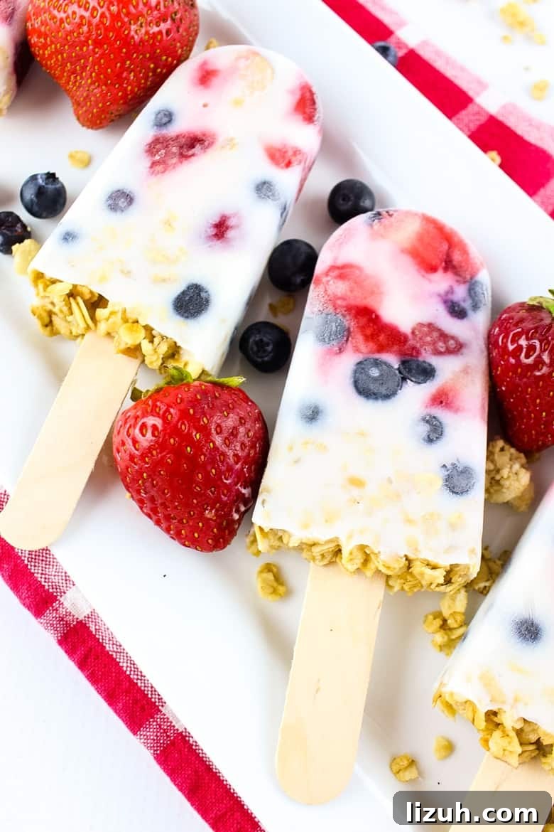 Creamy Yogurt Breakfast Popsicles with Fresh Fruit 6 Yogurt, berry, and granola popsicles on a white platter, with fresh strawberries and blueberries