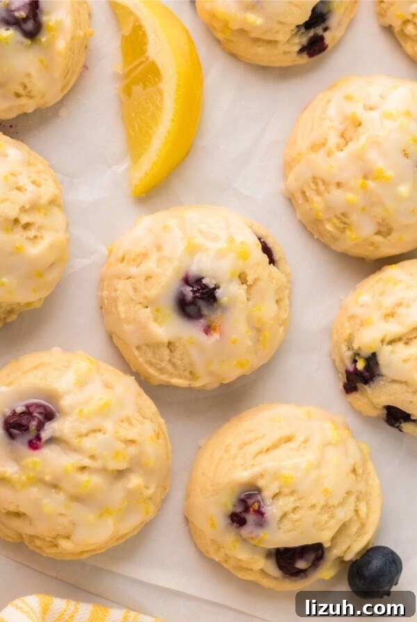 Lemon blueberry cookies on parchment paper with lemon wedges and blueberries scattered next to them.
