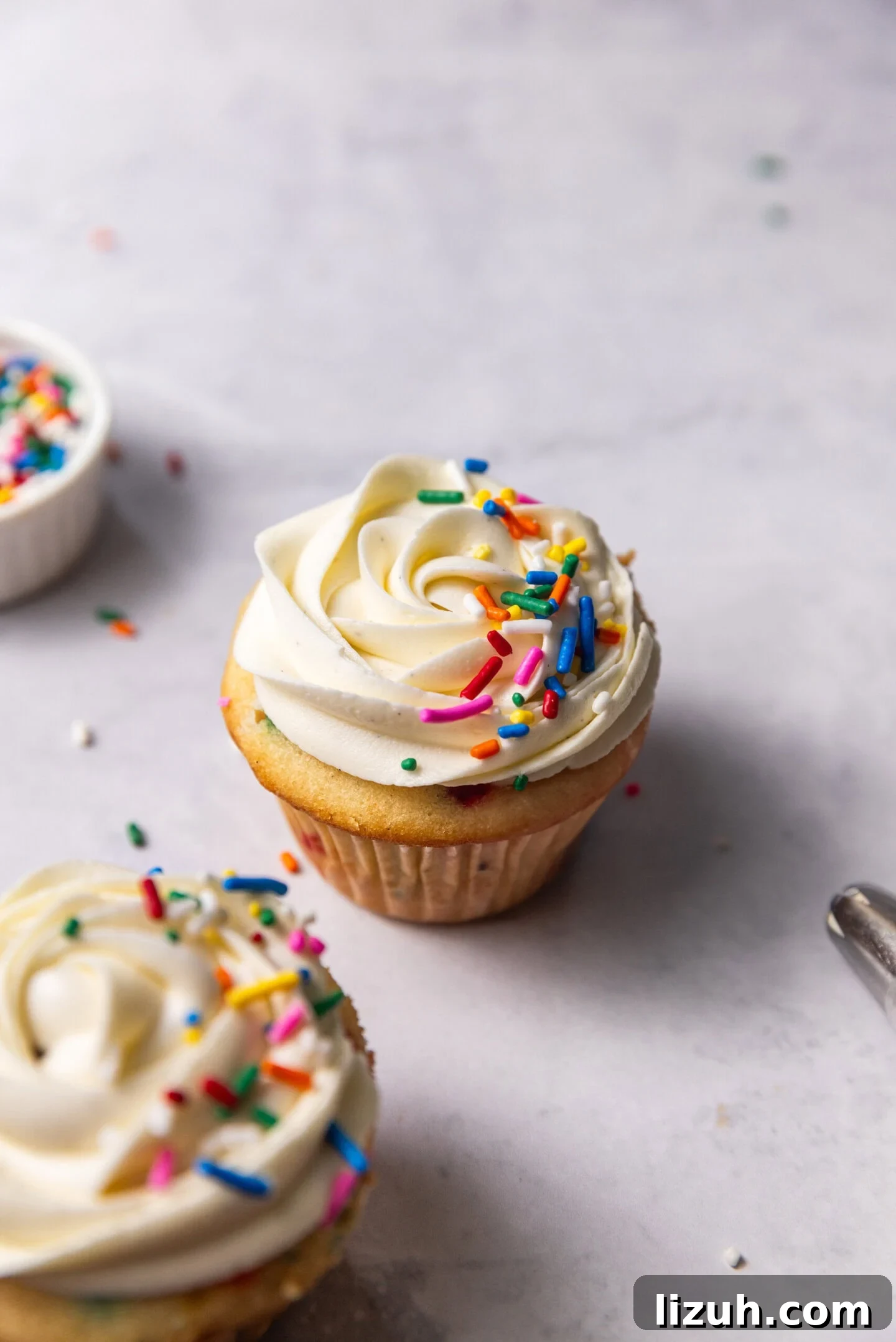 Two funfetti cupcakes with vanilla buttercream frosting and rainbow sprinkles on top sitting next to a small pinch pot of sprinkles and a piping bag filled with frosting.