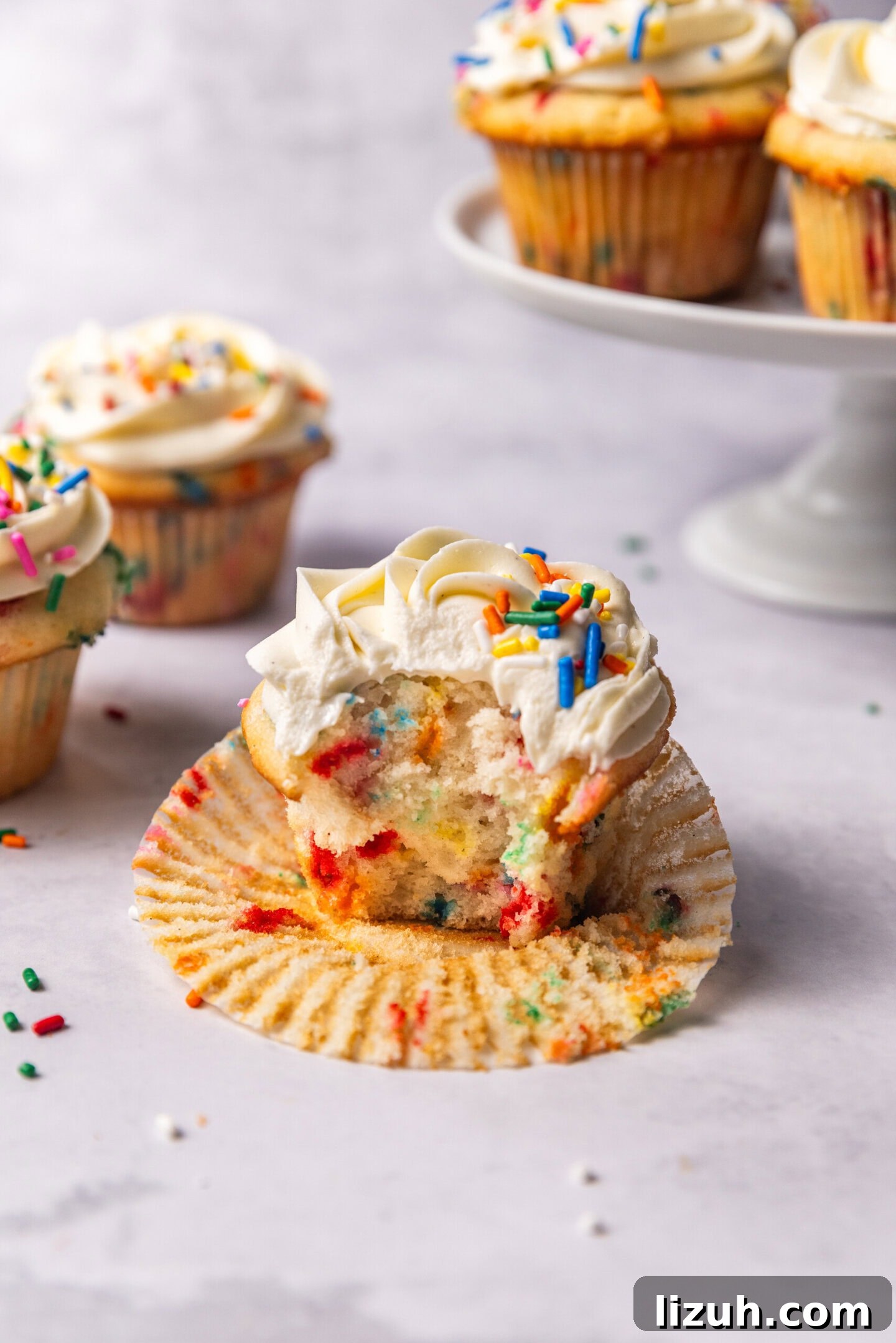 A bite taken out of a cupcake showing the moist, soft and fluffy interior with pops of color from the rainbow sprinkles. Other funfetti cupcakes are sitting in the background on top of a cake stand.