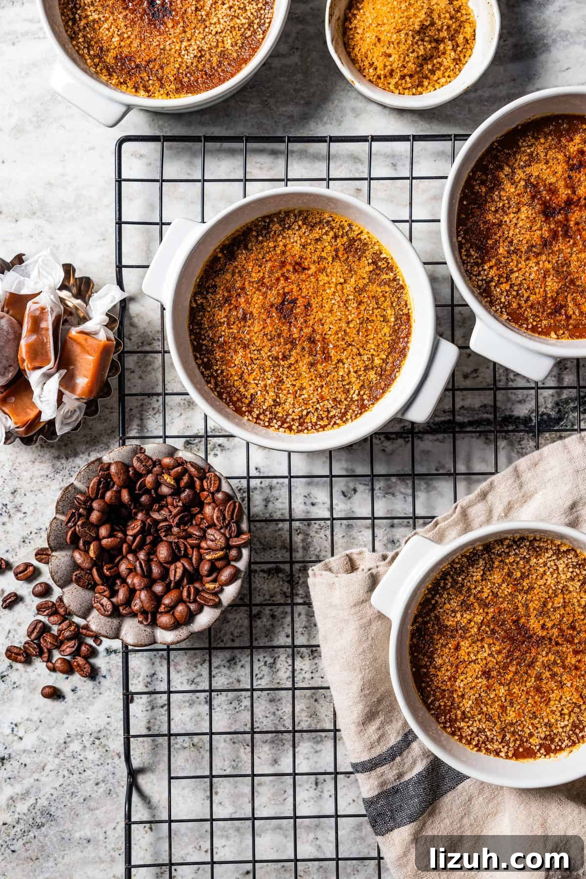 Crème brûlée in ramekins next to small bowls of coffee beans and caramels on a wire rack.