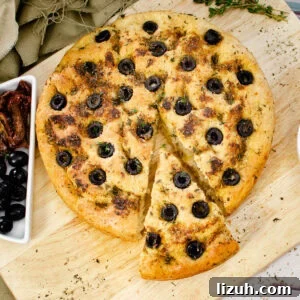 Olive and Herb Focaccia Bread Recipe: Crispy, Flavorful Loaf 3 Olive Focaccia Bread on a wooden chopping board with slice cut out served with black olives and sun dried tomatoes on the side