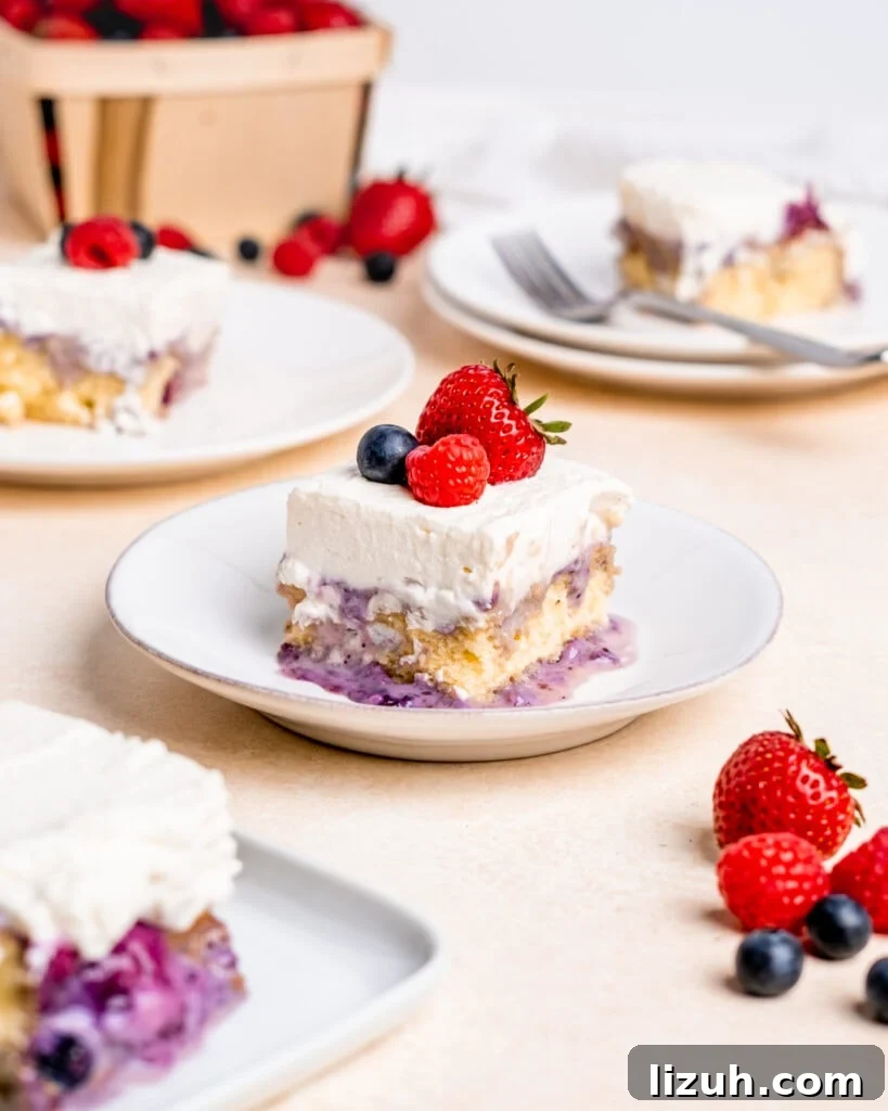 A slice of milk and berries cake on a white plate with a strawberry, raspberry, and blueberry on top of the whipped cream frosting.