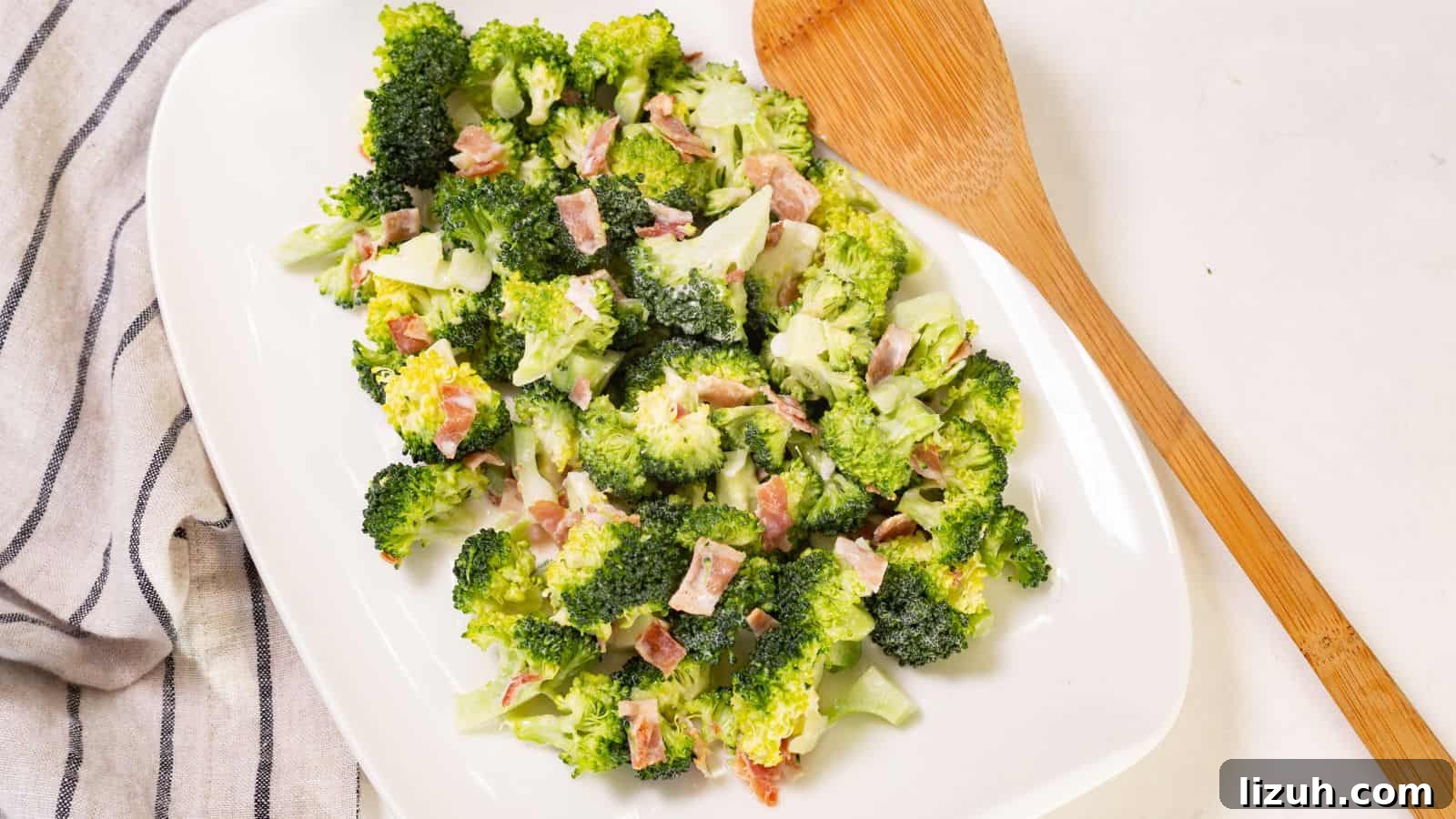 Broccoli salad with bacon bits on a white oval plate, accompanied by a wooden serving spoon on a linen napkin.