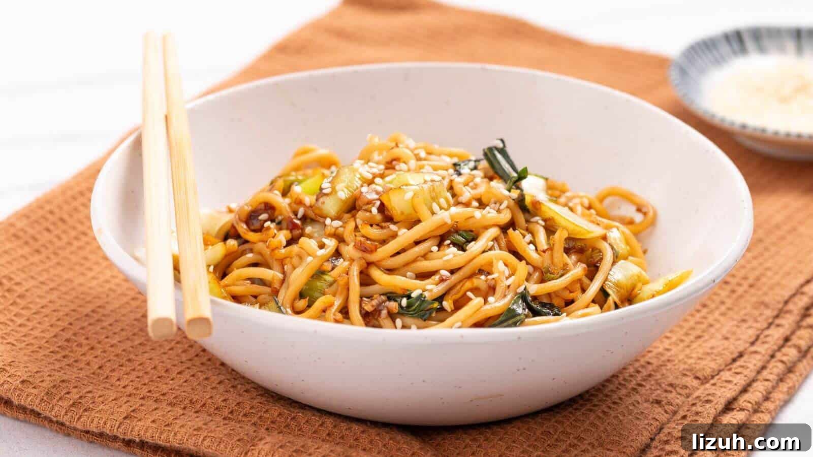 A bowl of stir-fried noodles with vegetables, topped with sesame seeds, placed on a brown cloth napkin. Chopsticks rest on the bowl's edge, and a small dish is visible in the background.