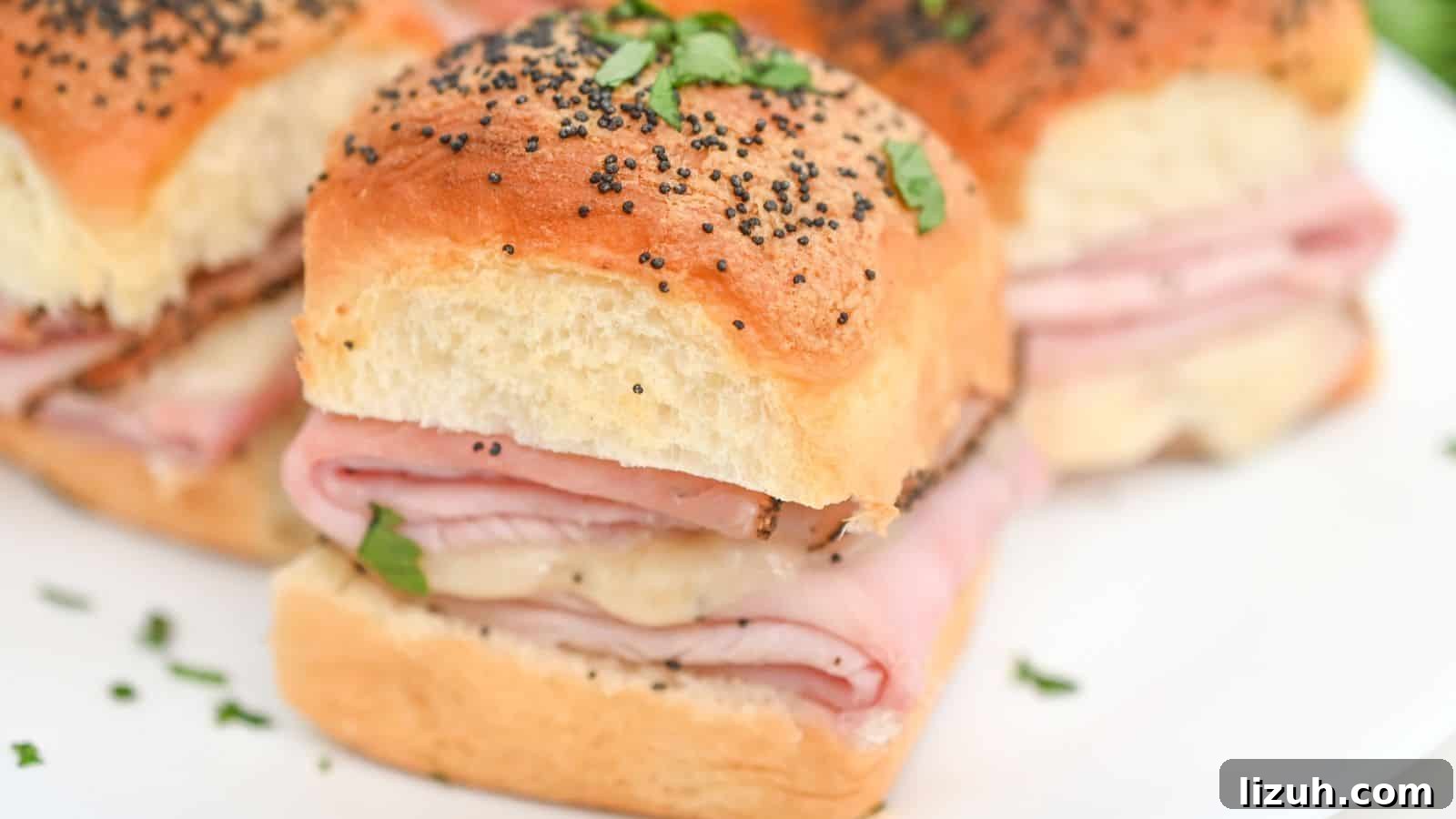 A close-up image of a platter of ham and cheese slider sandwiches sprinkled with poppy seeds and garnished with chopped herbs.