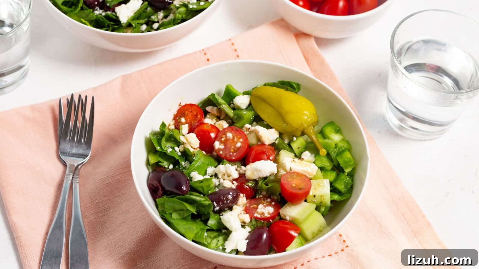 A fresh greek salad with tomatoes, feta cheese, cucumbers, olives, and peppers, served with a fork and a glass of water on the side.