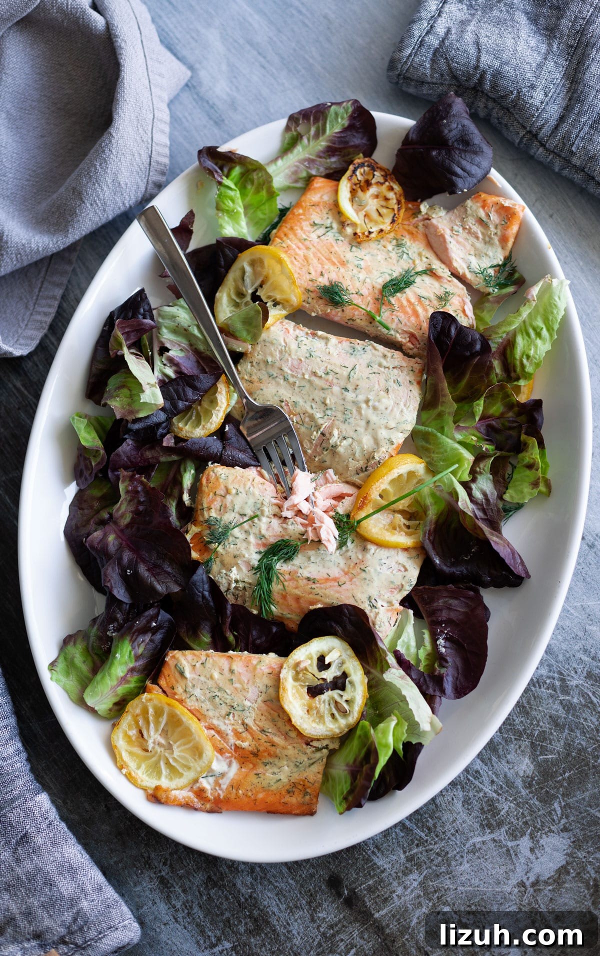 Salmon with dill sauce on a serving platter over a bed of mixed greens with roasted lemons.