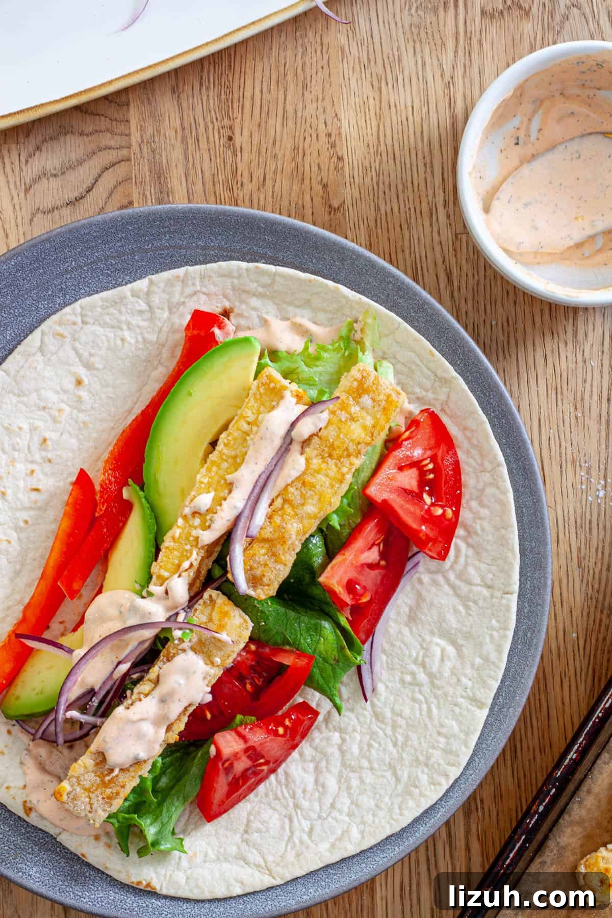 Crispy Buffalo Tofu Wraps with Cooling Ranch Slaw 11 A plate with crispy buffalo tofu wrap topped with avocado, red peppers, lettuce, red onions and a homemade buffalo ranch sauce.