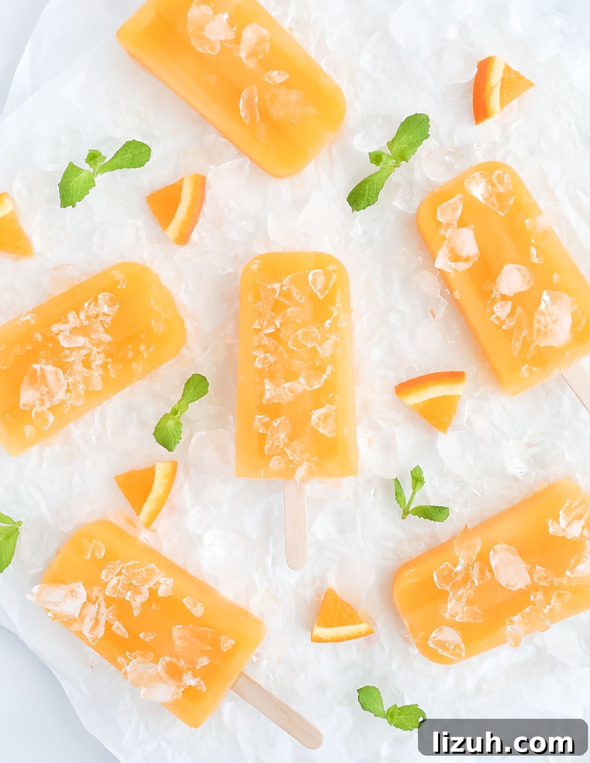 Homemade Orange Popsicles: Refreshing Citrus Ice Pops Recipe 2 Six orange ice pops on a sheet of white parchment paper, surrounded by crushed ice, fresh mint leaves, and orange slices