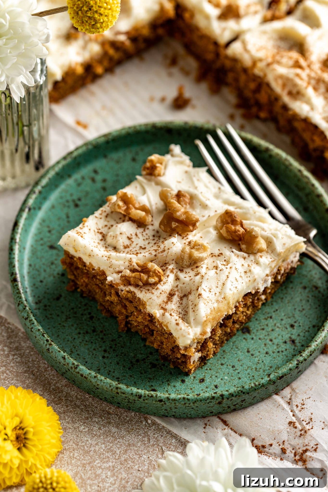 Chewy Carrot Cake Bars with Cream Cheese Frosting Recipe 2 Single slice of carrot cake bar on a rustic green plate with a fork, topped with frosting, walnuts, and cinnamon, with flowers in the background.