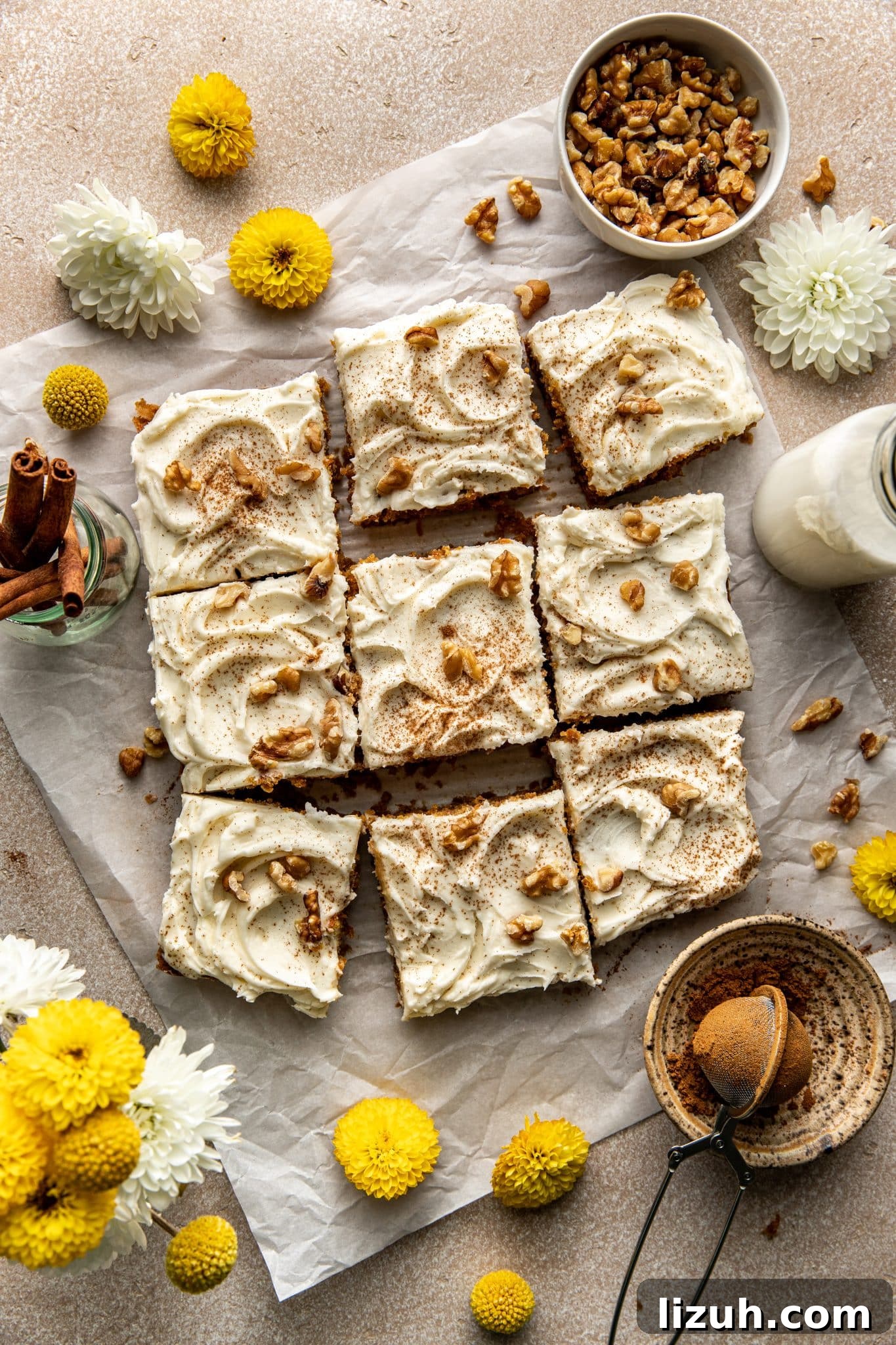Chewy Carrot Cake Bars with Cream Cheese Frosting Recipe 6 Carrot cake bars cut into squares on parchment paper, topped with cream cheese frosting, cinnamon, and chopped walnuts, surrounded by fresh flowers and cinnamon sticks.