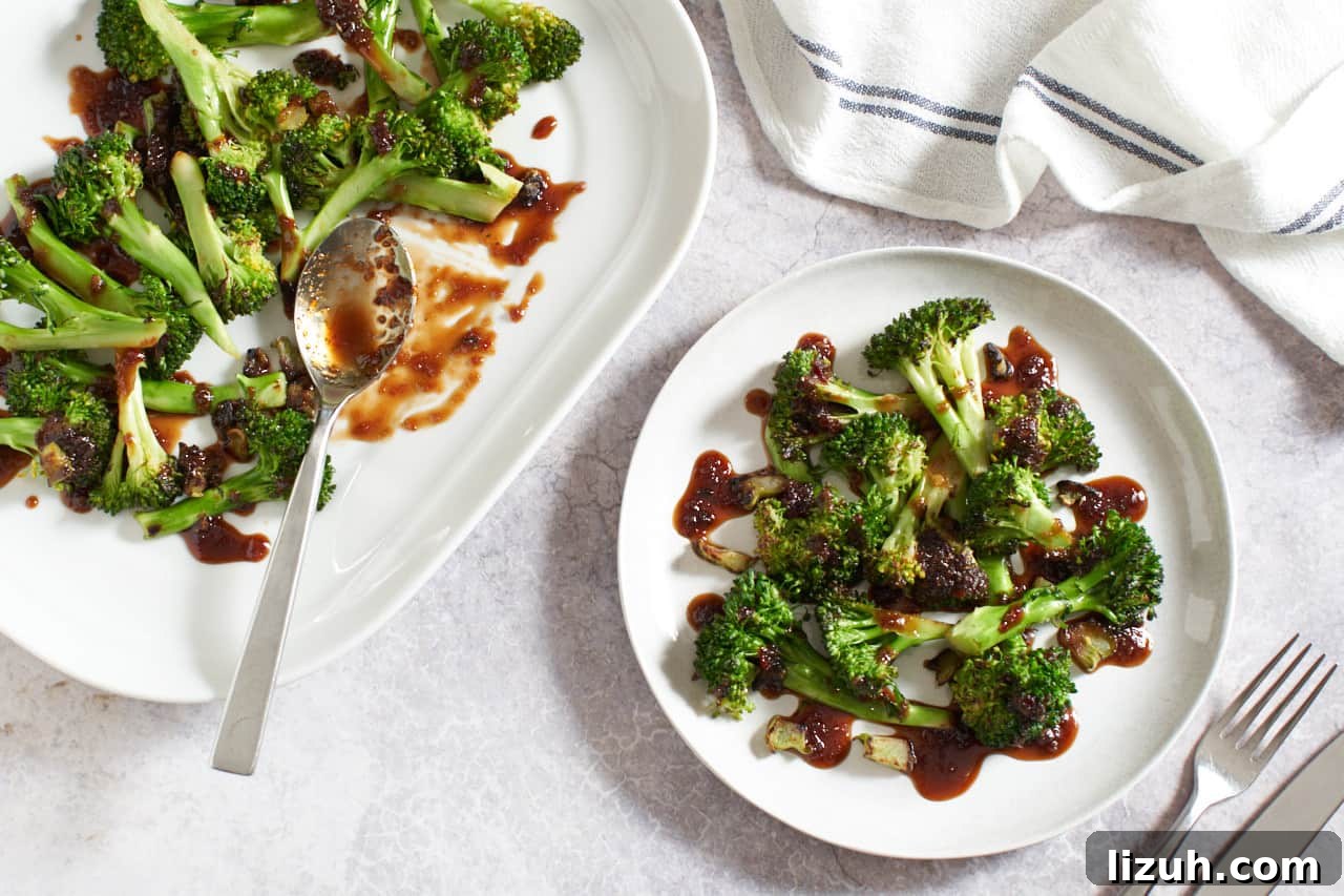 A platter of spicy Asian broccoli with a spoon on it next to a plate of the same. A white and blue napkin is on the upper right, utensils are on the lower right.