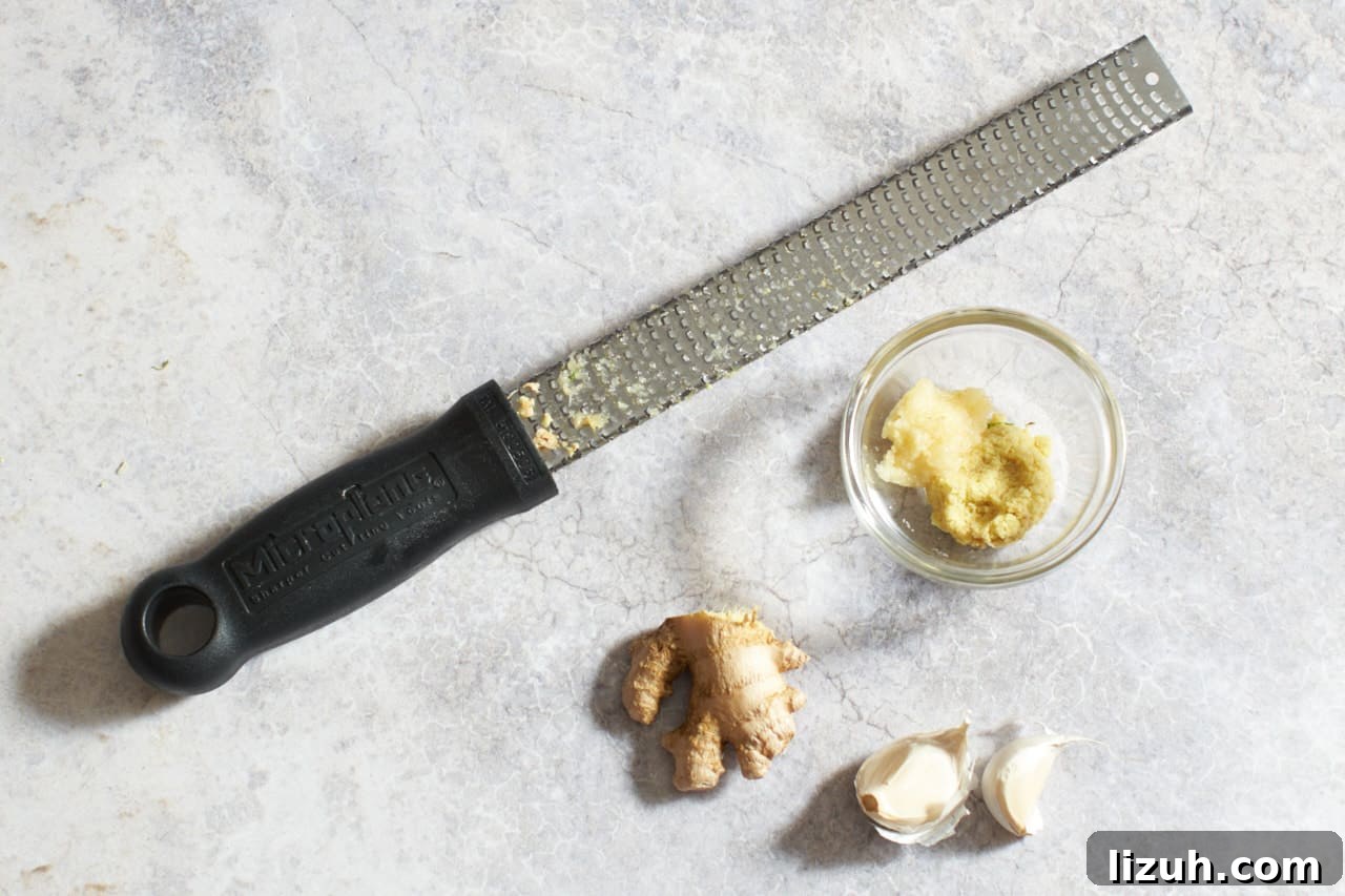 A Microplane grater next to garlic cloves, fresh ginger, and a small glass bowl of grated ginger and garlic.