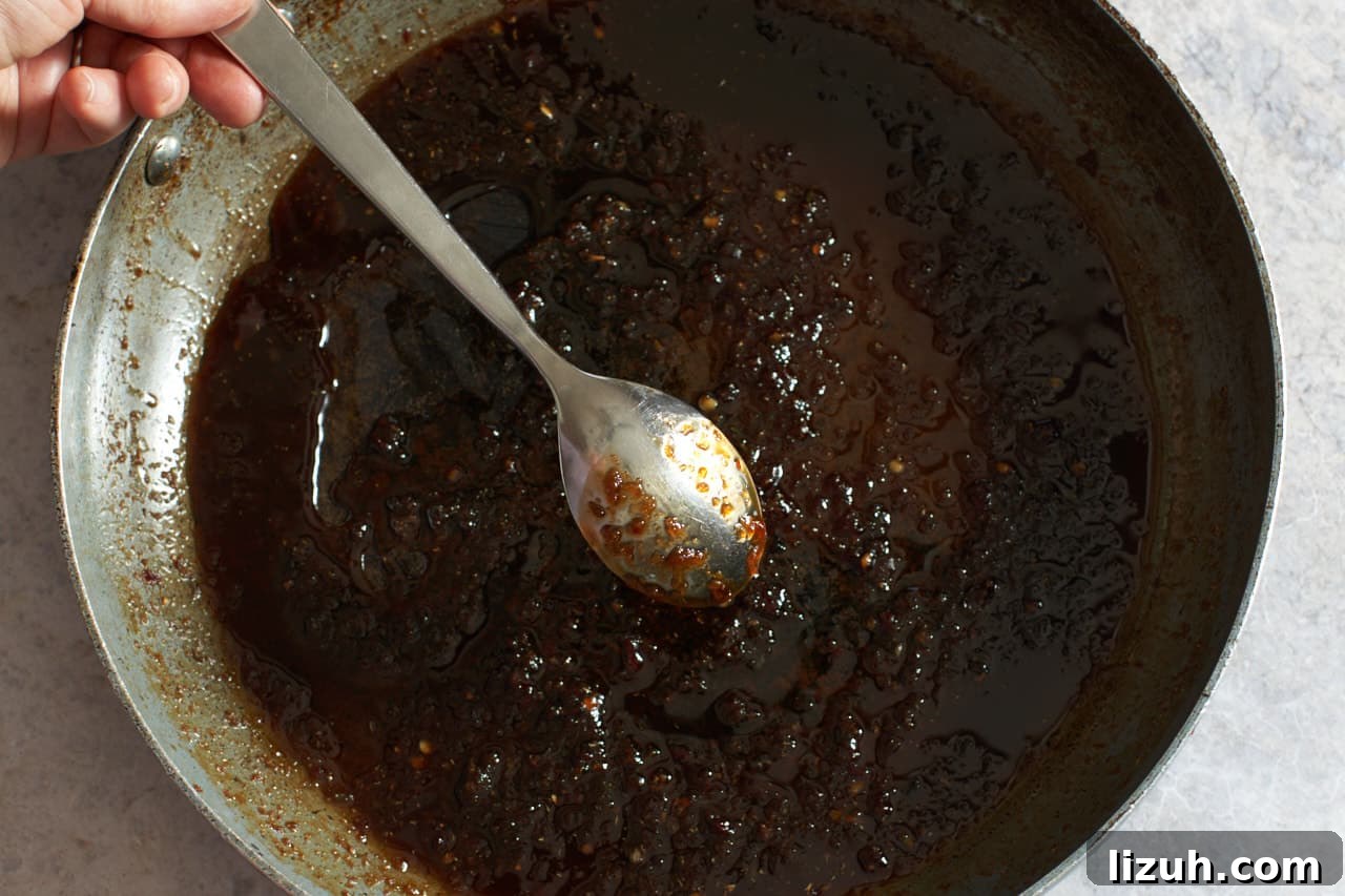 A woman's hand holds a spoon coated with spicy sauce over a skillet to show how thick the sauce is.