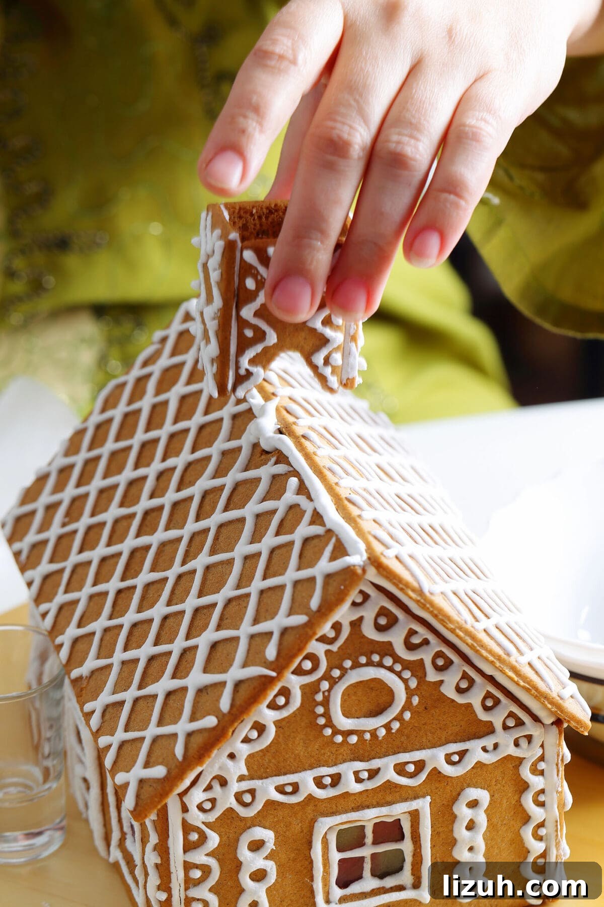 Host a Festive Gingerbread House Party: Tips, Themes & Checklist 5 a hand presses a gingerbread chimney on top of a decorated gingerbread house with white icing as the decoration