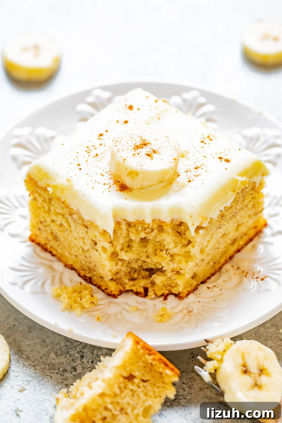 A slice of banana cake with cream cheese icing on a plate with a corner bite missing. 