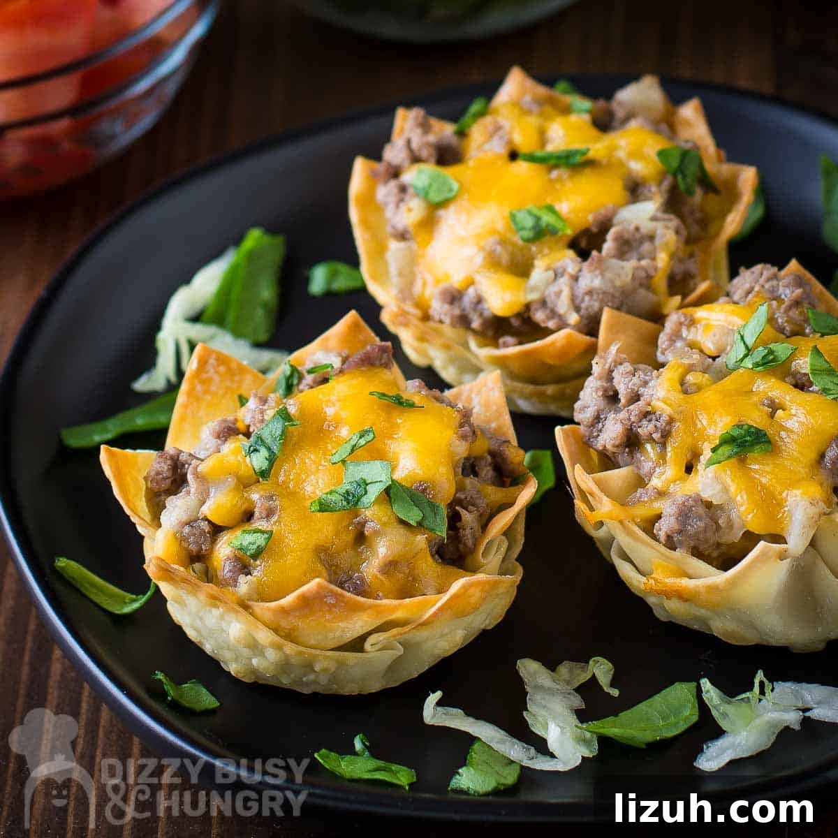 Crispy Wonton Cheeseburger Cups — Bite-Sized Party Sliders 7 Side shot of three wonton cheeseburger cups on a black plate garnished with herbs on a wooden surface.