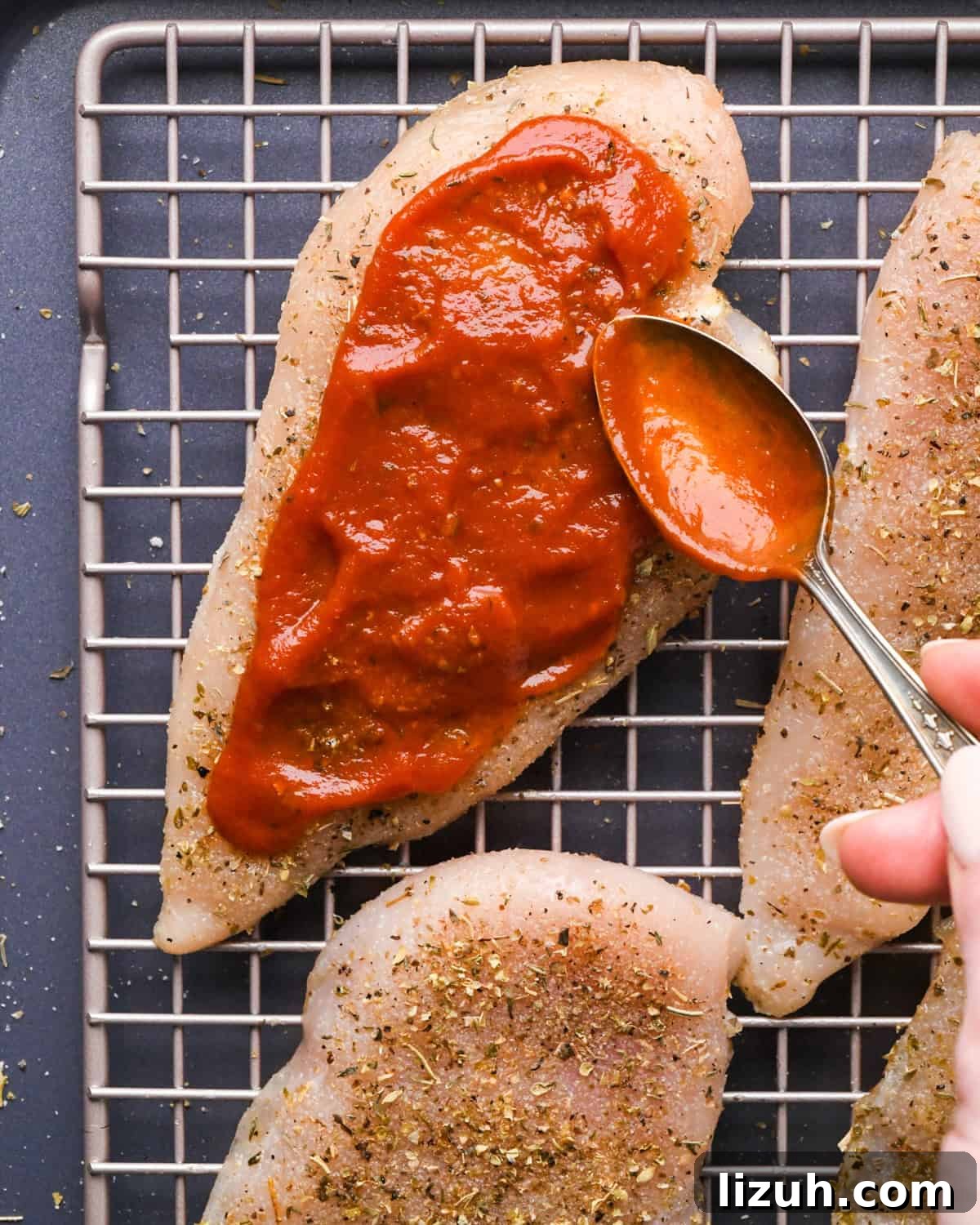 Making pizza chicken: close-up photo of chicken breasts sprinkled with spices and pizza sauce being spread on top.